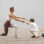 A woman and a boy exercise together on a wooden deck by the lake, promoting healthy lifestyle.