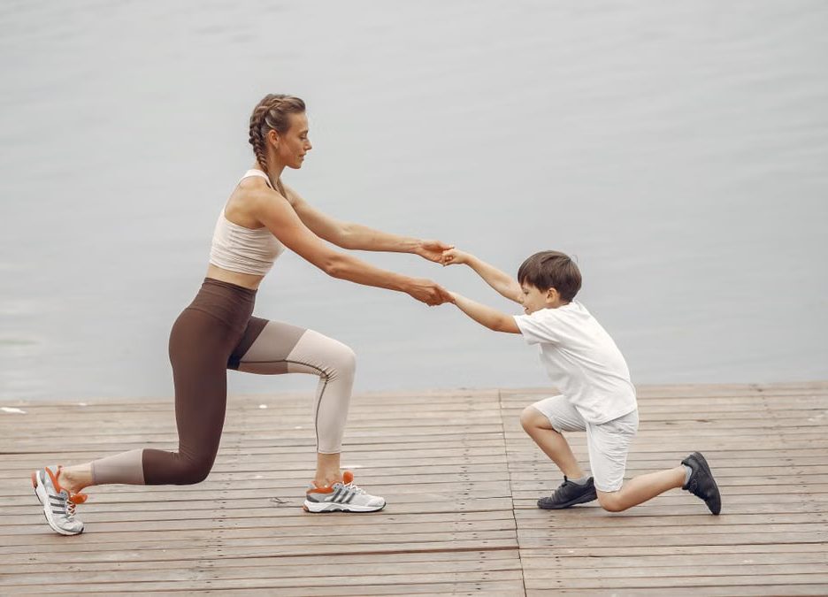 A woman and a boy exercise together on a wooden deck by the lake, promoting healthy lifestyle.
