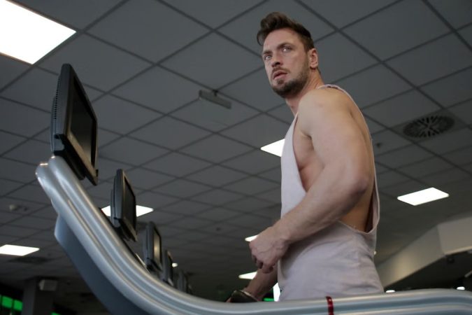Low angle of muscular adult male athlete in sportswear during training on running machine in modern spacious fitness center while looking away