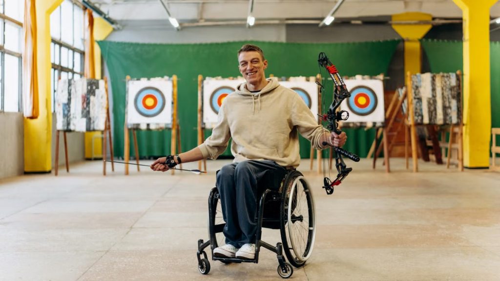 A disabled archer smiles while holding a bow in an indoor archery range.