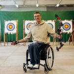 A disabled archer smiles while holding a bow in an indoor archery range.