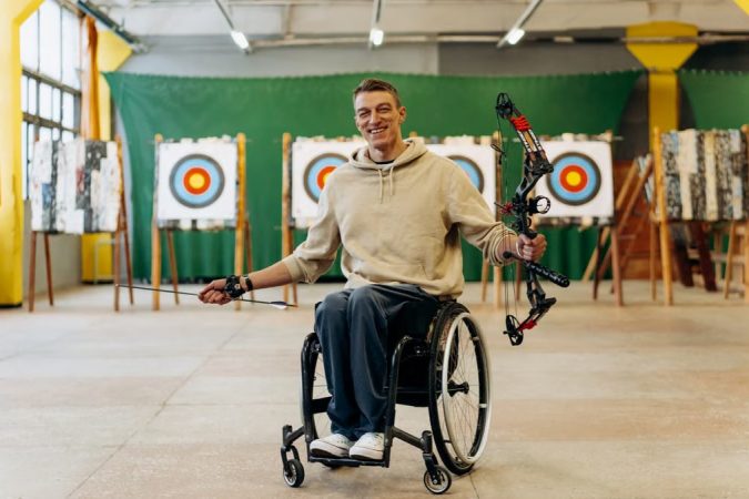 A disabled archer smiles while holding a bow in an indoor archery range.