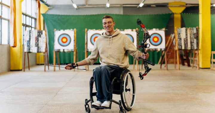 A disabled archer smiles while holding a bow in an indoor archery range.