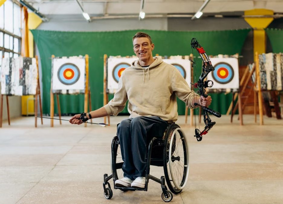 A disabled archer smiles while holding a bow in an indoor archery range.